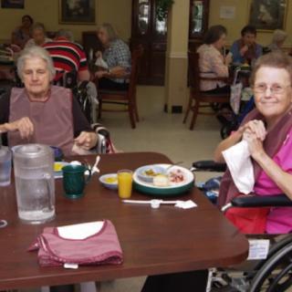 Two seniors seated at a dining table, enjoying a meal in a nursing home environment.