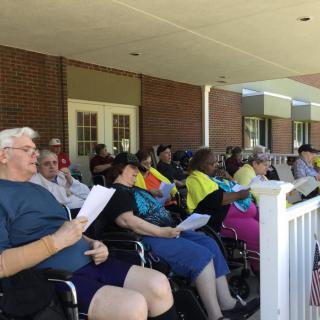 A group of people seated on a porch, some in wheelchairs, engaging in a reading activity.