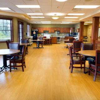 A spacious dining area with wooden flooring, multiple tables and chairs, and a kitchen in the background.
