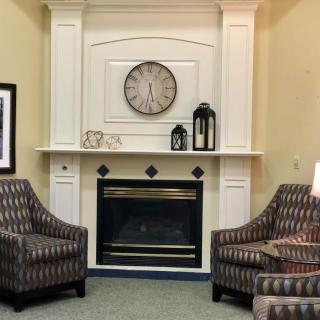 A cozy living room with a white fireplace, adorned with a clock and lanterns, flanked by patterned armchairs.