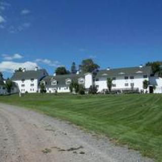 A long white building with multiple sections, surrounded by a well-manicured lawn and trees under a clear blue sky.