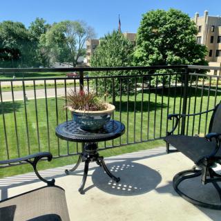 A sunny balcony with outdoor furniture, featuring a table with a potted plant and views of lush greenery.