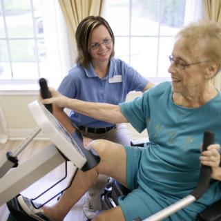 An elderly woman works out on a fitness machine under the guidance of a healthcare professional.