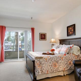 A cozy bedroom with a neatly made bed, decorative pillows, and a lamp on the nightstand. Red curtains frame the window leading to a balcony.