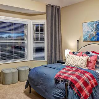 A cozy, well-decorated bedroom featuring a neatly made bed with colorful pillows and a plaid blanket, soft lighting, and a large window with blinds.