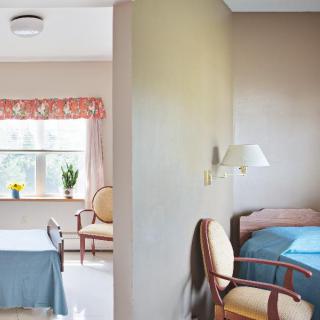 A cozy interior with a bedroom and living area, featuring chairs, tables, and natural light from a window.