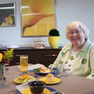 An elderly woman smiling while seated at a table with a meal in a yellow and green themed dining area.
