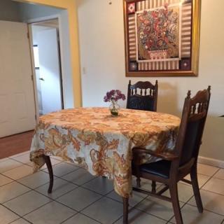 A cozy dining area with a table covered in a floral tablecloth, flanked by two wooden chairs and adorned with a vase of flowers. A decorative artwork hangs on the wall.