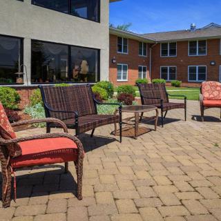 A sunny outdoor patio area with wicker chairs and tables on a brick surface, adjacent to a building.