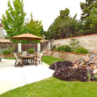 A well-maintained backyard with a patio set under an umbrella, surrounded by lush landscaping and a green lawn.