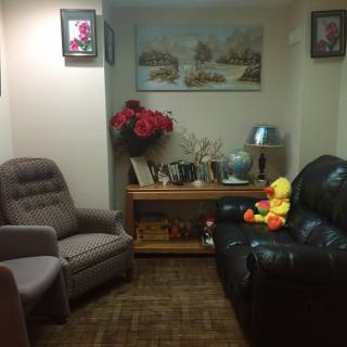 A cozy living room with a sofa and armchair, decorated with paintings, flowers, and a stuffed toy.