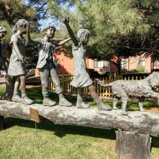 A bronze sculpture of children and a dog balancing on a log in a park setting.
