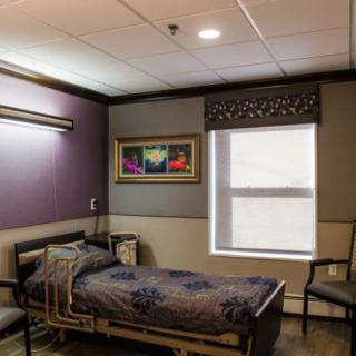A hospital room featuring a single bed, chairs, a painting on the wall, and a window with blinds.