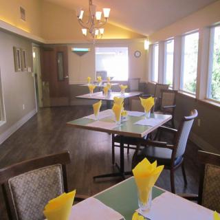 A cozy dining room with tables set for a meal, featuring yellow napkins and ample natural light from large windows.