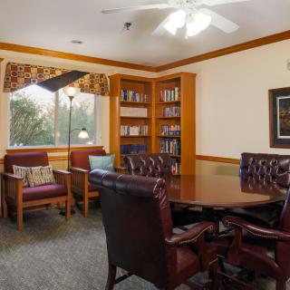 A cozy library room with a central table surrounded by chairs, bookshelves, and a window view.