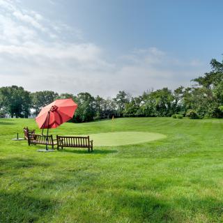 A scenic golf course with a green, bench, and red umbrella under a clear sky.