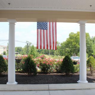 An American flag hanging between two white columns on a patio with greenery in the background.
