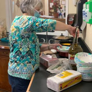 A woman in a colorful shirt prepares food in a kitchen while wearing a mask.
