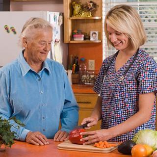 A caregiver assists an elderly person in the kitchen, chopping vegetables together.