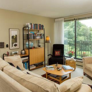 A cozy living room with a comfortable sofa, bookshelf, coffee table, and a large window providing a view of greenery.