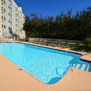An outdoor swimming pool next to a building, surrounded by trees under a clear blue sky.