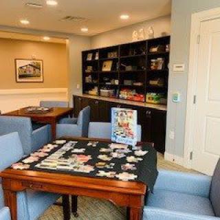 A cozy library space featuring a puzzle in progress on a wooden table surrounded by comfortable seating and bookshelves filled with various items.