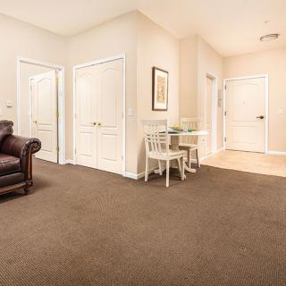 A cozy living room with beige walls, a brown carpet, and white doors. The space includes a leather sofa and a small table with two chairs.