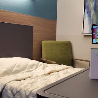 A cozy hotel room featuring a neatly made bed, a green chair, and a workspace with a screen.