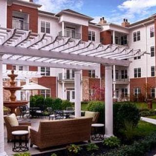 A serene outdoor patio with a wooden pergola, featuring comfortable seating and a fountain, surrounded by well-maintained landscaping.