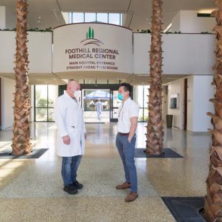 Two individuals, one in a lab coat, converse in the lobby of a medical center with palm trees and an open entrance.