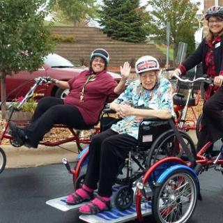 A group of people enjoying a ride outdoors with bicycles adapted for wheelchairs, all wearing helmets and smiling.