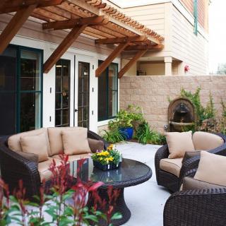 A cozy patio with comfortable outdoor furniture under a pergola, featuring a small fountain and various plants.
