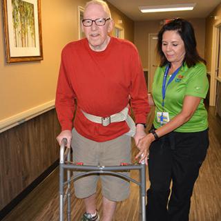 An elderly man using a walker is assisted by a caregiver in a hallway.