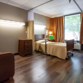 A well-lit hospital room with a single bed, wooden flooring, and large windows.