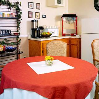 A cozy kitchen setup featuring a table with a red tablecloth, chairs, and various appliances including a microwave, popcorn machine, and coffee maker.