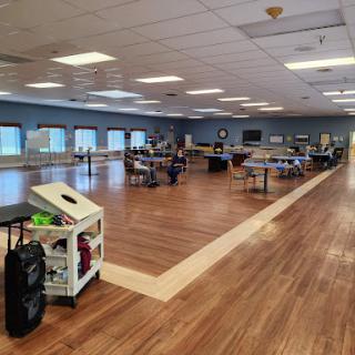 A spacious community room featuring wooden floors and a bright, organized layout with tables and chairs set up for activities.