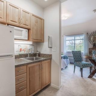 A compact kitchen in an apartment features wooden cabinets, a microwave, and a refrigerator opening into a cozy dining area with large windows.
