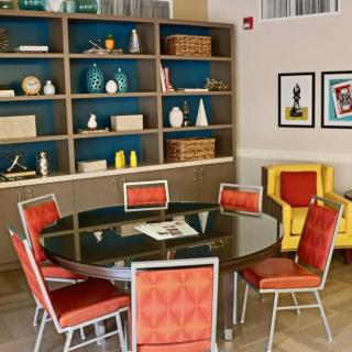 A stylish interior with a round glass table surrounded by red chairs, adjacent to a bookshelf filled with decorative items.