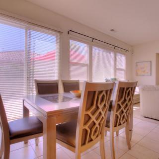 A bright dining room with a wooden table and chairs, large windows, and blinds.