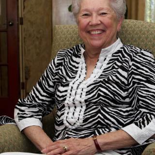 A smiling elderly woman sitting indoors wearing a zebra pattern shirt.