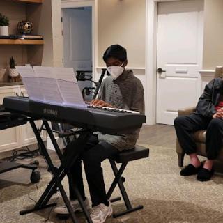 A young musician performs on a digital piano while wearing a mask, with an audience member seated nearby.