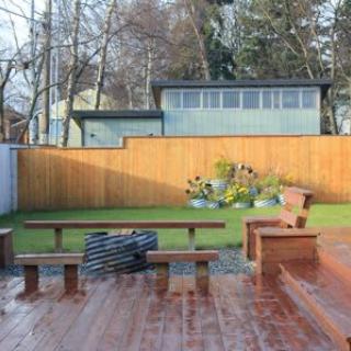 A well-maintained backyard featuring a wooden deck with benches, a green lawn, and a garden with plants in the corner.