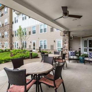 An outdoor patio area with tables and chairs adjacent to an apartment building, featuring landscaping and a ceiling fan.