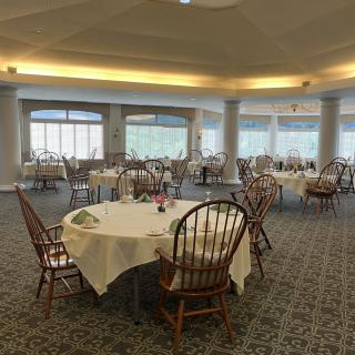 An empty dining room with round tables and chairs, featuring large windows and a carpeted floor.