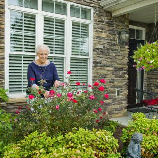 An elderly woman tending to her garden in front of a brick house.
