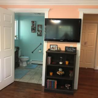 A cozy living space featuring a wall-mounted television, a bookshelf stocked with books and decorative items, and a glimpse into an adjacent bathroom.