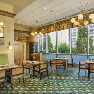 A modern restaurant interior with stylish decor, featuring tables and chairs under elegant hanging lights, set against large windows with a view of greenery.
