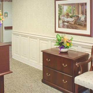 An office reception area with a desk, cabinet, chair, and decorative elements like flowers and a painting.