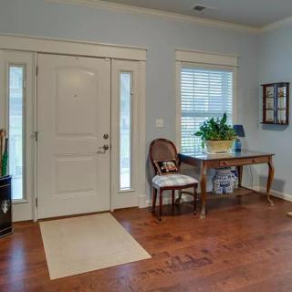 A welcoming home entrance with a hardwood floor, featuring a front door flanked by two narrow windows. A decorative table with a plant and lamp adds charm, accompanied by an elegant chair.