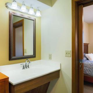 A cozy bathroom with a mirror, sink, and overhead lighting adjacent to a bedroom.